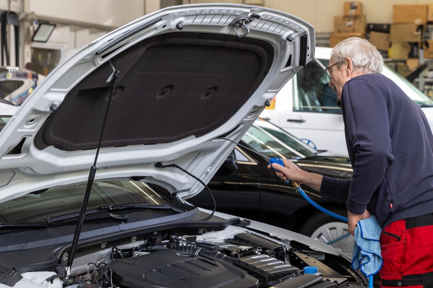 BlitzSauber das Reinigungszentrum Rellingen UG - Fahrzeugpflege - Älterer Mann reinigt den Motorraum eines weißen Autos mit einem blauen Luftgebläse und einem blauen Tuch in einer Garage