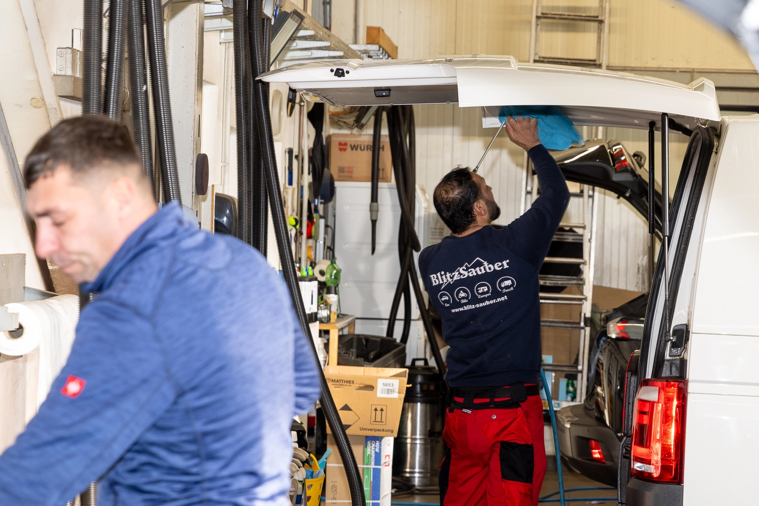 BlitzSauber das Reinigungszentrum Rellingen UG - Fahrzeugpflege - Zwei Männer arbeiten in einer Werkstatt; einer reinigt die Innenseite einer Fahrzeughecktür, während der andere im Vordergrund verschwommen zu sehen ist.