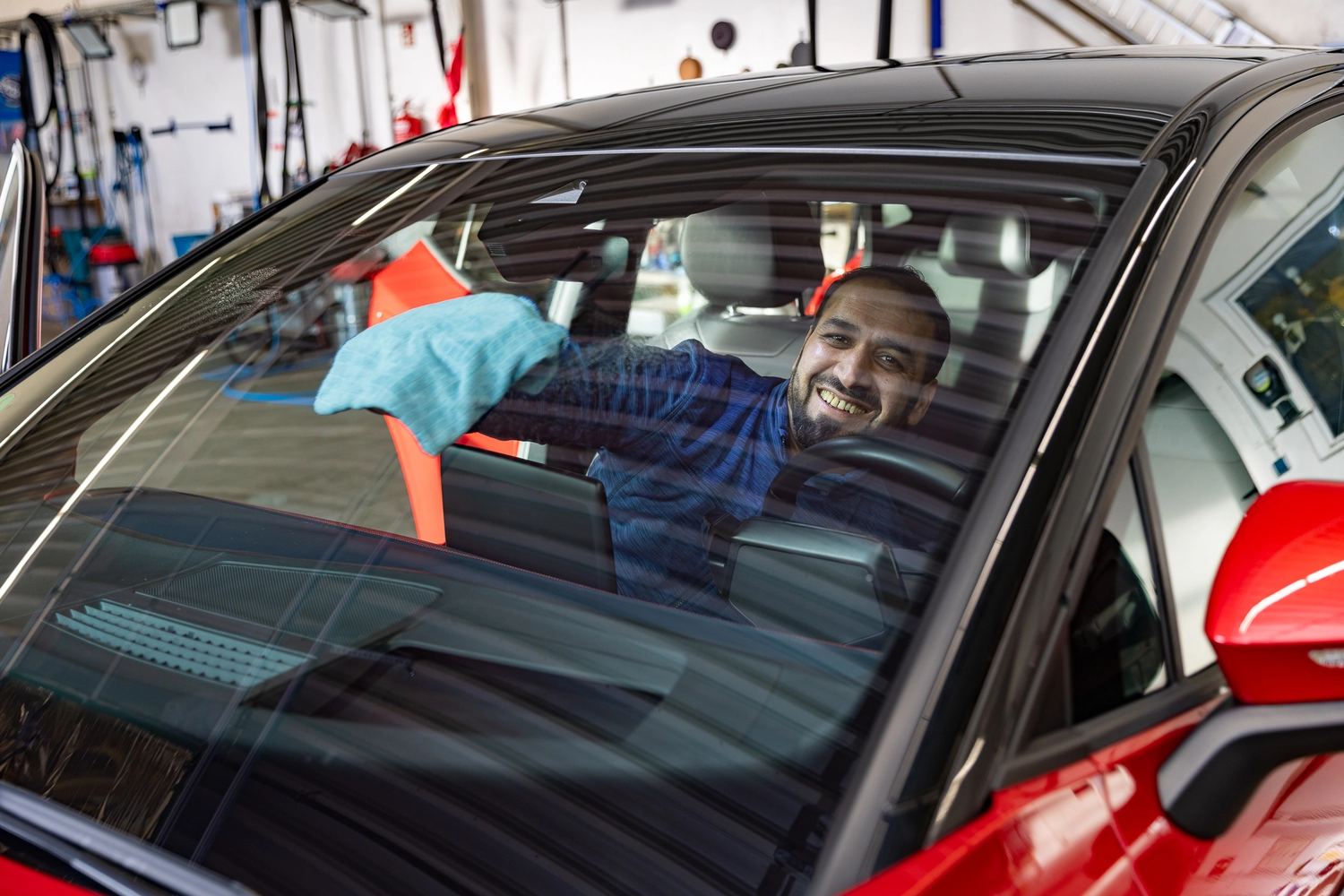 BlitzSauber das Reinigungszentrum Rellingen UG - Fahrzeugpflege - Lächelnder Mann in einem roten Auto, der mit einem blauen Tuch die Windschutzscheibe reinigt