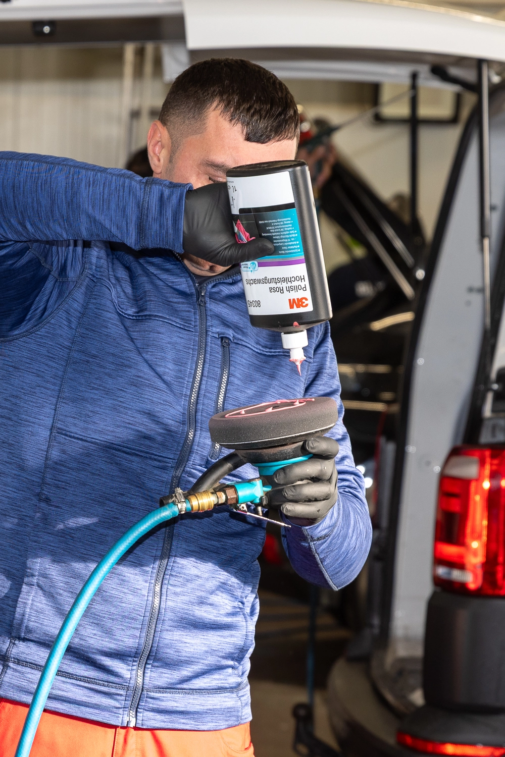 BlitzSauber das Reinigungszentrum Rellingen UG - Fahrzeugpflege - Person in blauer Jacke und schwarzen Handschuhen, die in einer Garage 3M-Politur auf einen Autopuffer aufträgt