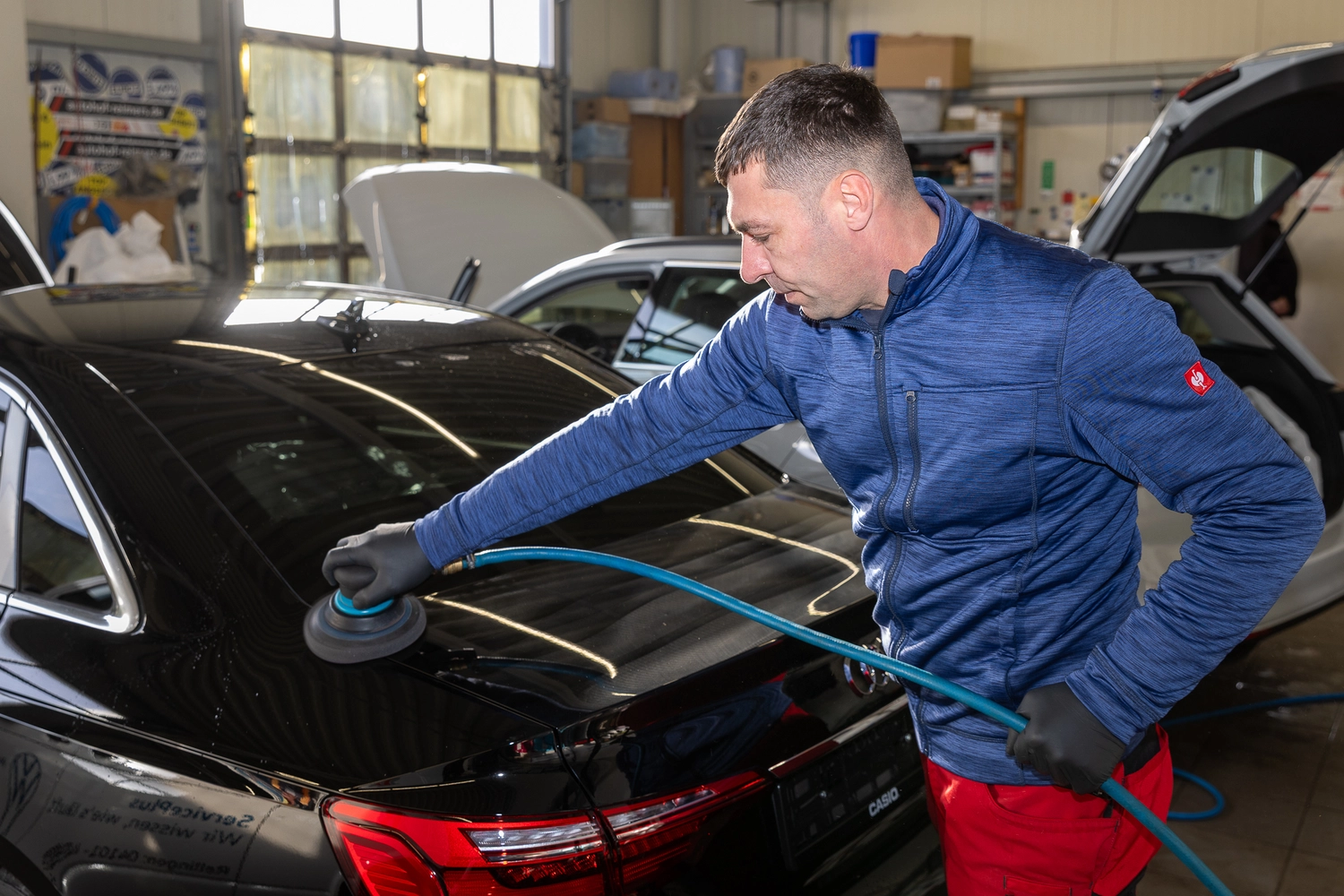 BlitzSauber das Reinigungszentrum Rellingen UG - Fahrzeugpflege - Mann in blauer Jacke und roter Hose poliert in einer Garage den Kofferraum eines schwarzen Autos mit einem Rotationspuffer