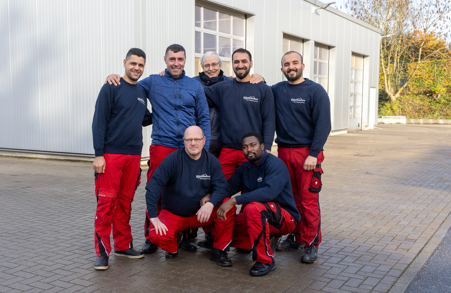 BlitzSauber das Reinigungszentrum Rellingen UG - Fahrzeugpflege - Sieben Männer posieren vor einem hellgrauen Industriegebäude, in roten Hosen und marineblauen oder blauen Jacken, teilweise mit „Blitzsauber“-Logo