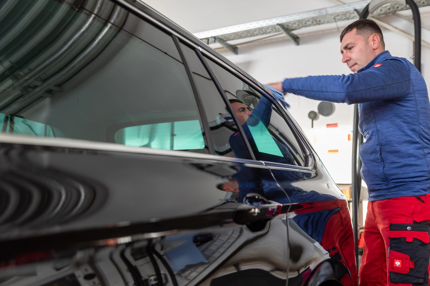 BlitzSauber das Reinigungszentrum Rellingen UG - Fahrzeugpflege - Mann in blauer Jacke und roter Hose reinigt in einer Garage die Scheibe eines schwarzen Autos mit einem Tuch