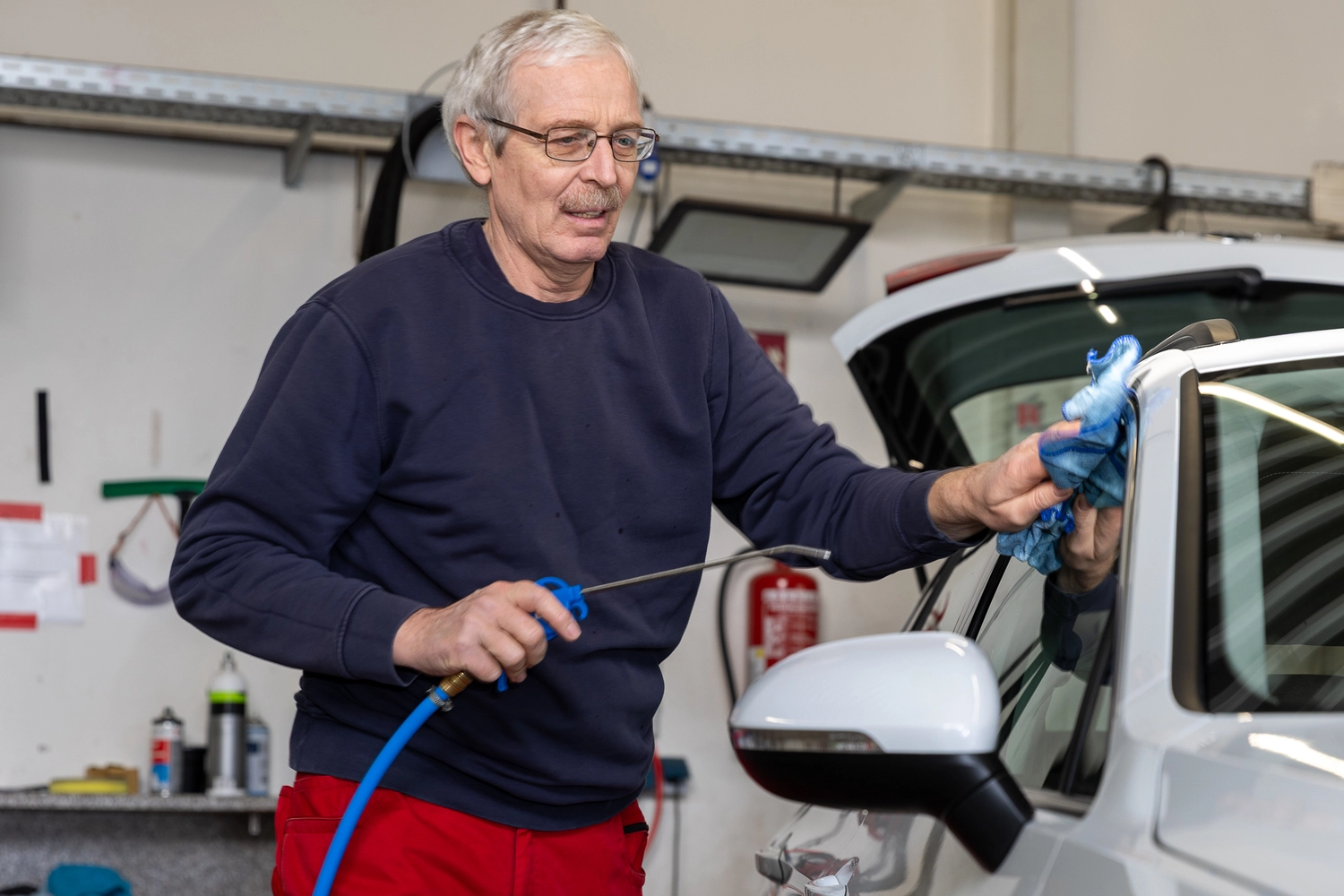 BlitzSauber das Reinigungszentrum Rellingen UG - Fahrzeugpflege - Mann in dunkelblauem Sweatshirt und roter Hose reinigt im Innenbereich eine weiße Autoscheibe mit einem blauen Tuch und einer Sprühdüse