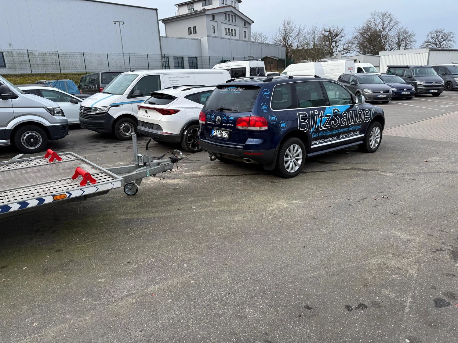 BlitzSauber das Reinigungszentrum Rellingen UG - Fahrzeugpflege - Auf einem Parkplatz geparkter blauer BlitzSauber-SUV, befestigt an einem Pkw-Anhänger aus Metall, umgeben von verschiedenen weißen und grauen Transportern und Autos