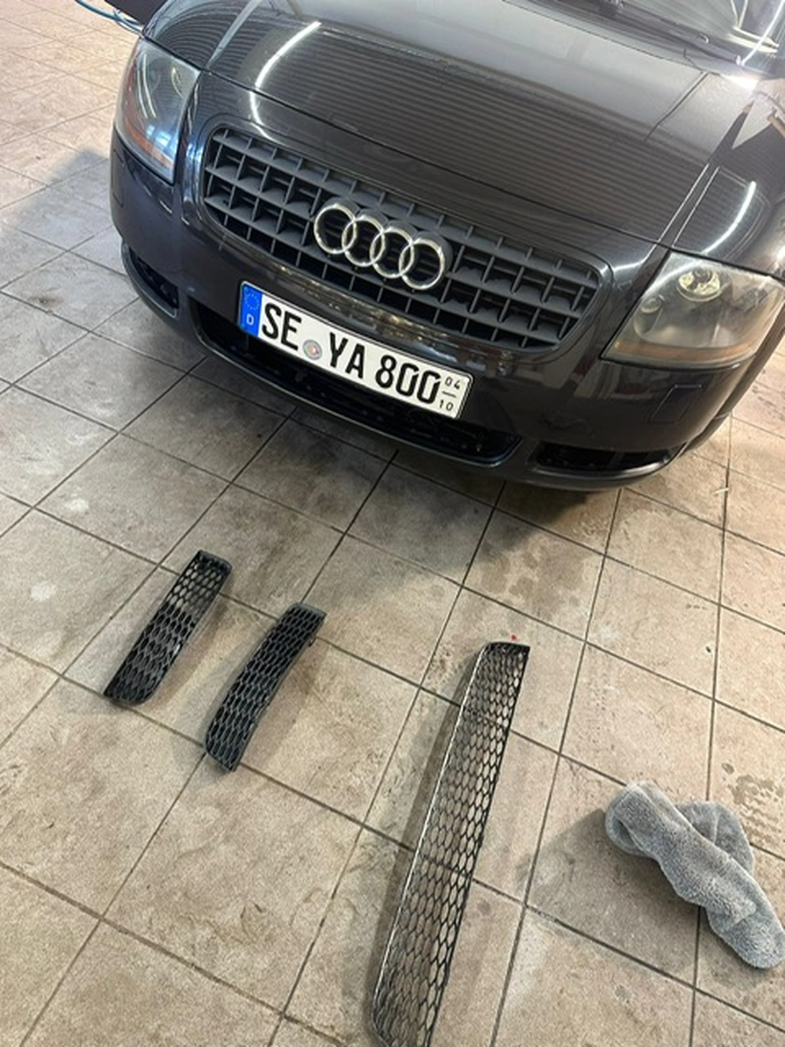 BlitzSauber das Reinigungszentrum Rellingen UG - Fahrzeugpflege - Frontansicht eines schwarzen Audi mit drei schwarzen Gittereinsätzen, die auf einem gefliesten Boden davor liegen, neben einem grauen Tuch