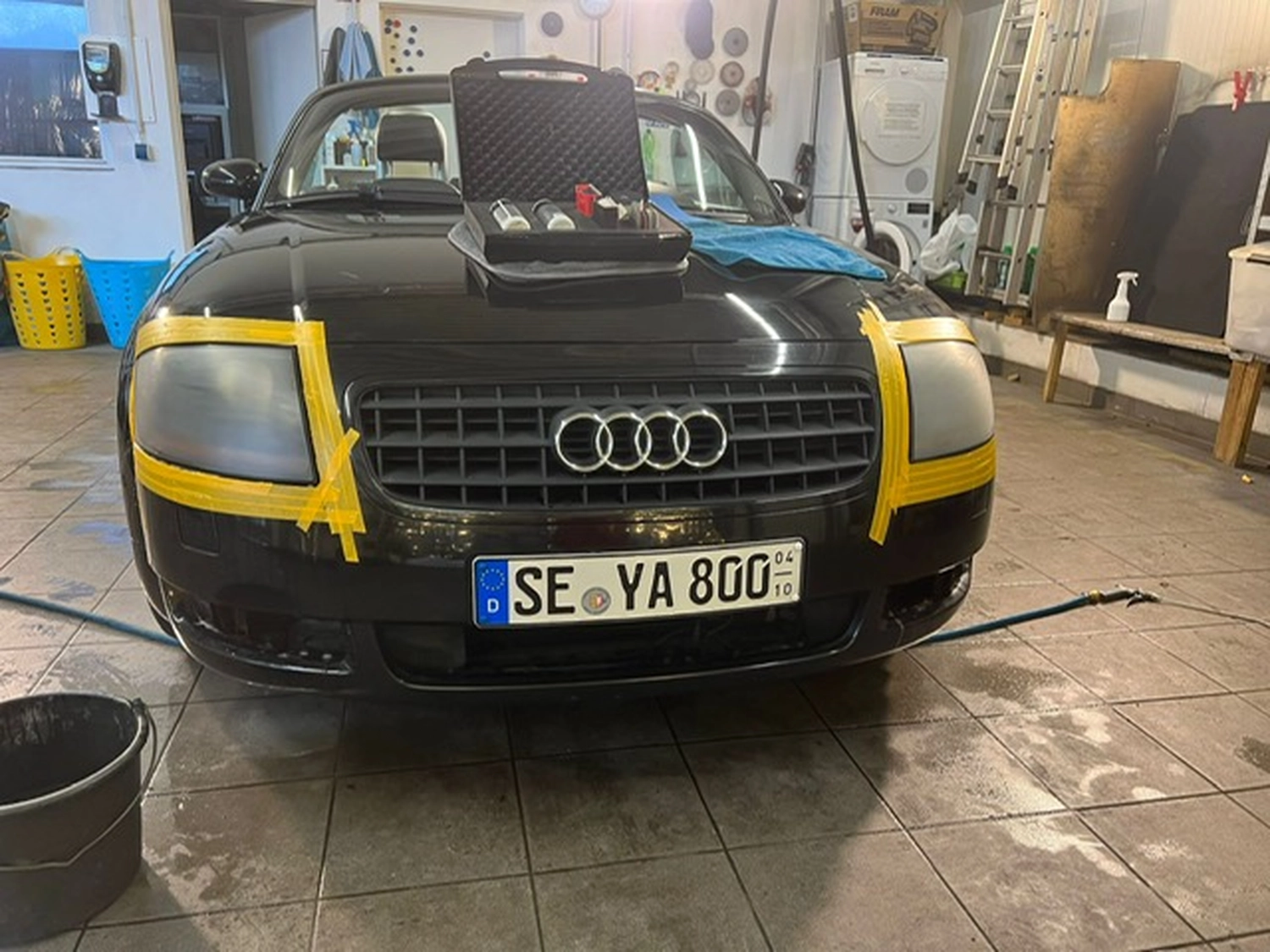 BlitzSauber das Reinigungszentrum Rellingen UG - Fahrzeugpflege - Schwarzer Audi in einer Garage mit durch gelbes Klebeband abgedeckten Scheinwerfern und Detaillierungszubehör auf der Motorhaube.
