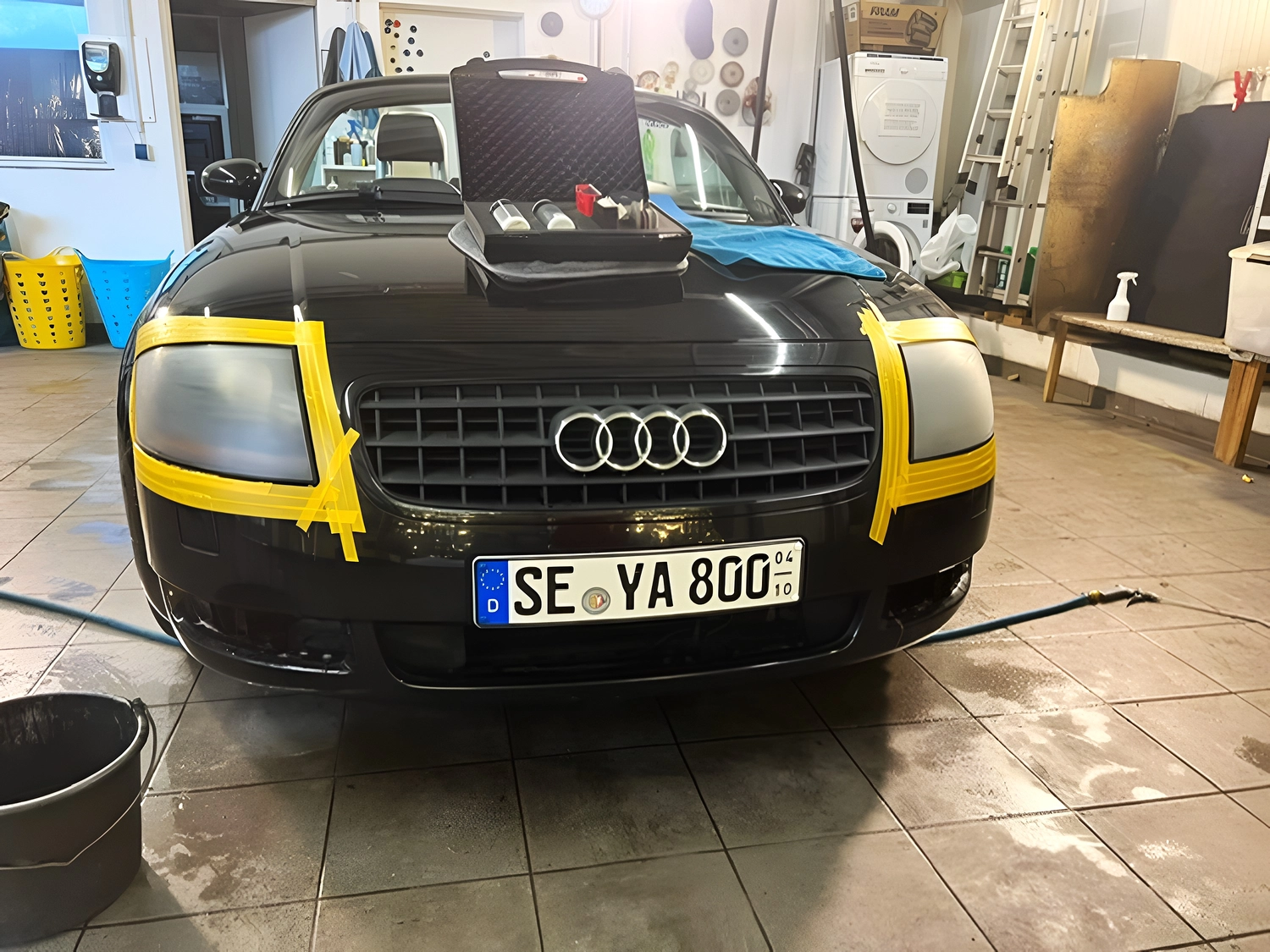 BlitzSauber das Reinigungszentrum Rellingen UG - Fahrzeugpflege - Schwarzer Audi in einer Garage mit durch gelbes Klebeband abgedeckten Scheinwerfern und Detaillierungszubehör auf der Motorhaube
